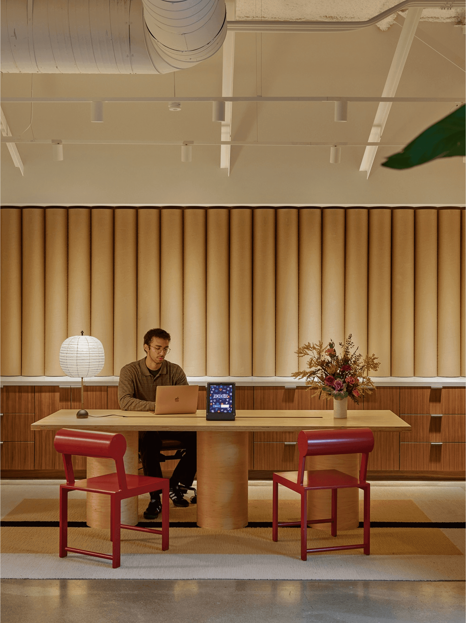 A man working on a laptop at a wooden desk with red chairs in a modern office setting
