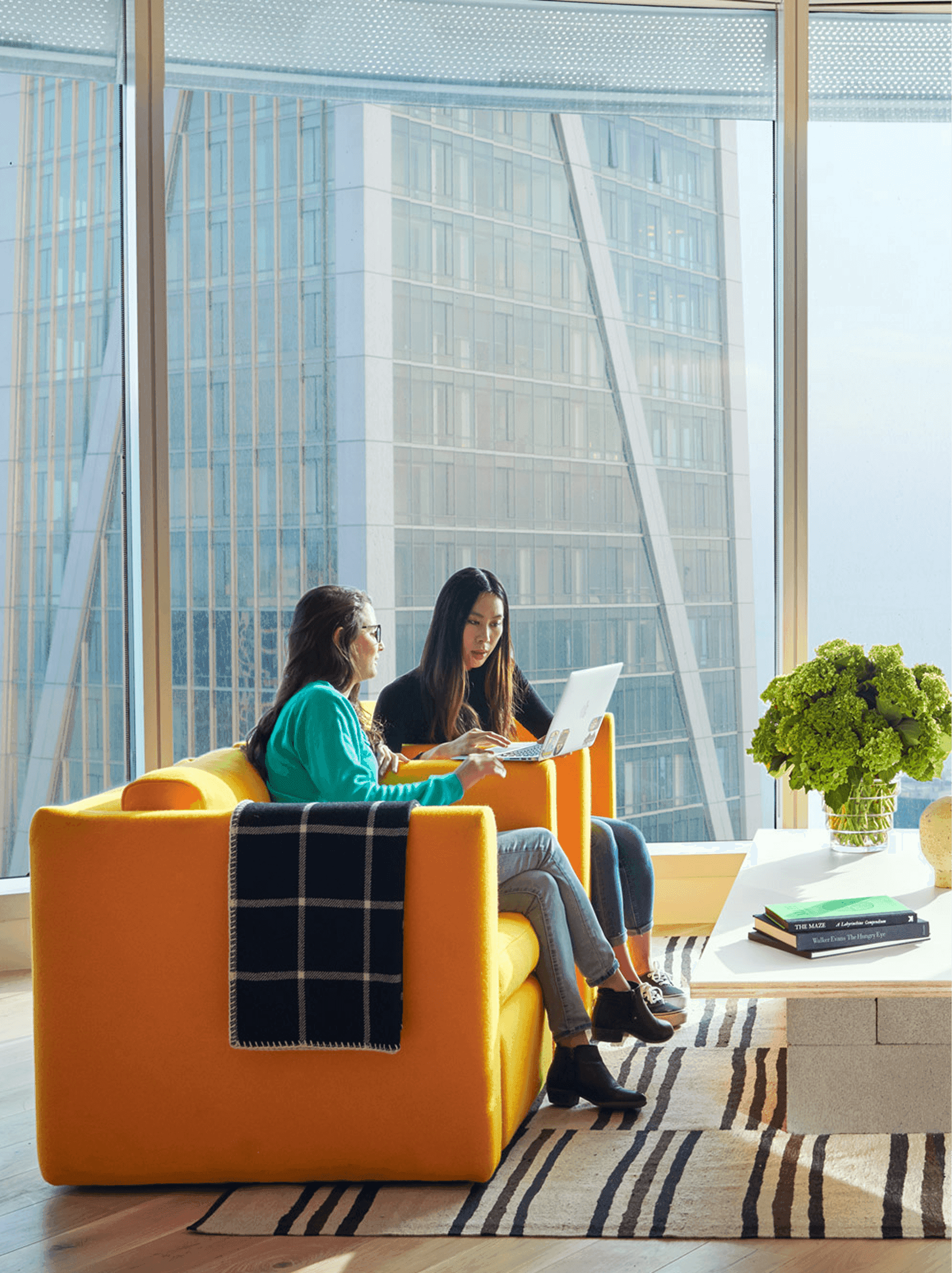 Two women collaborating on a laptop while seated on a yellow couch near a window