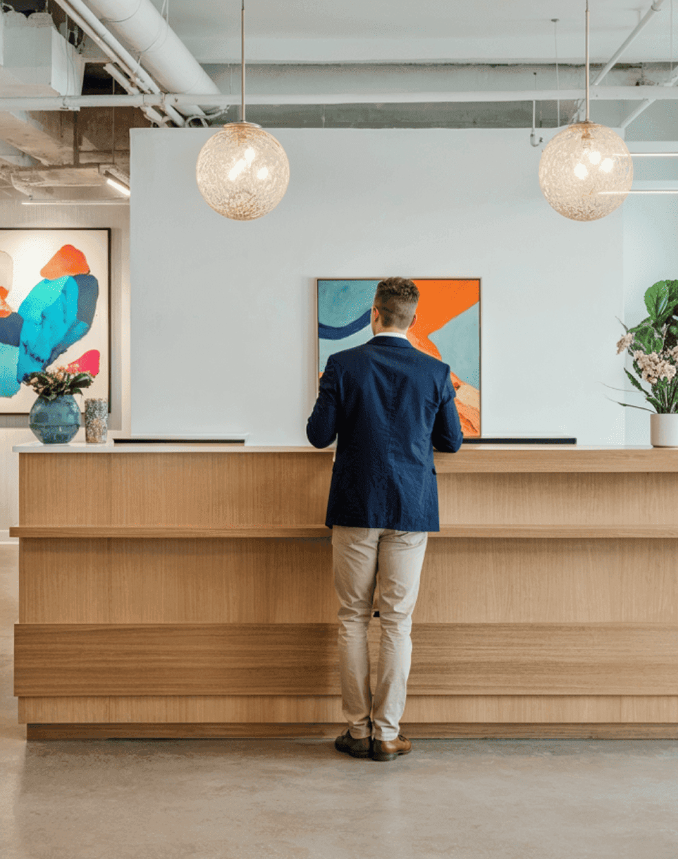 A man standing at a modern reception desk in an office, with abstract art and pendant lights in the background.