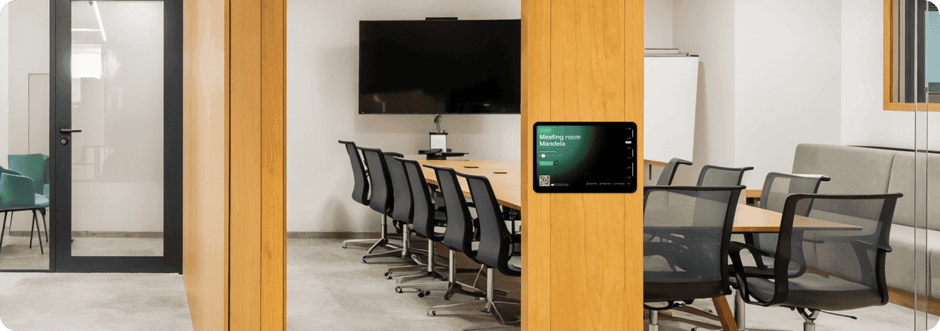 A modern meeting room with a large table, chairs, a TV screen, and a wall-mounted device, likely for access control or room booking.