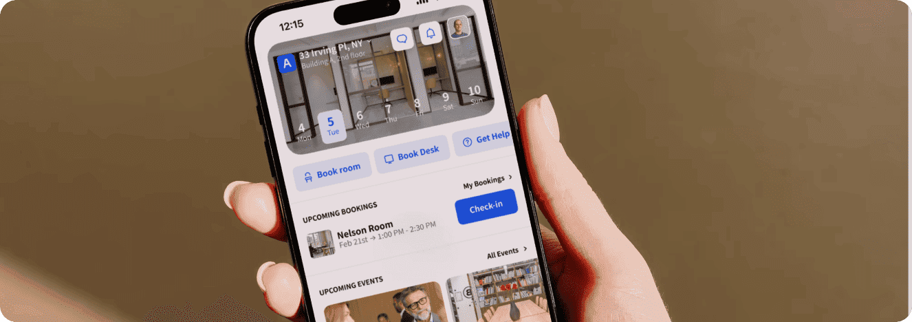 A hand holding a smartphone displaying a workspace booking app with options to book a room or desk, check in, and view upcoming bookings and events.