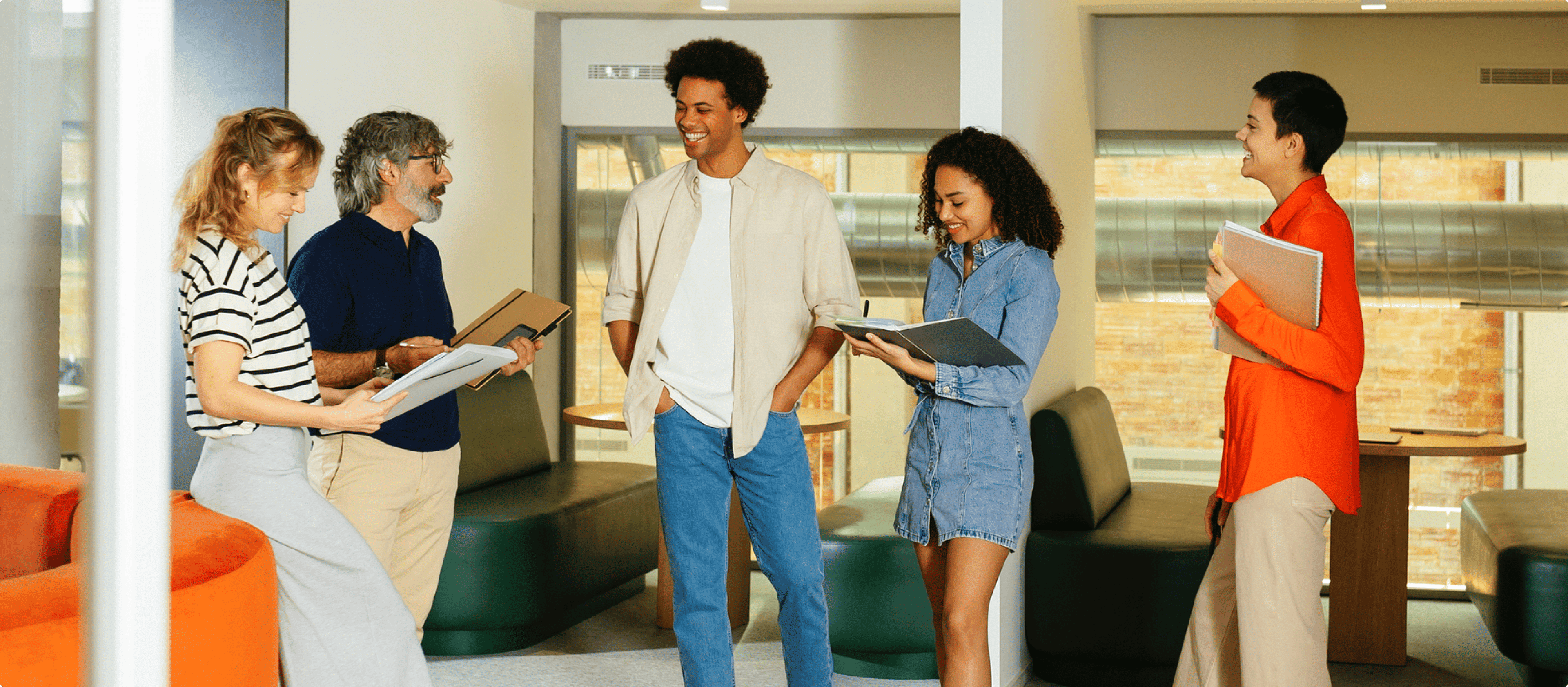 A group of five people in an office setting, engaged in conversation. Four individuals are holding notebooks or tablets, and they are standing near office chairs and desks.