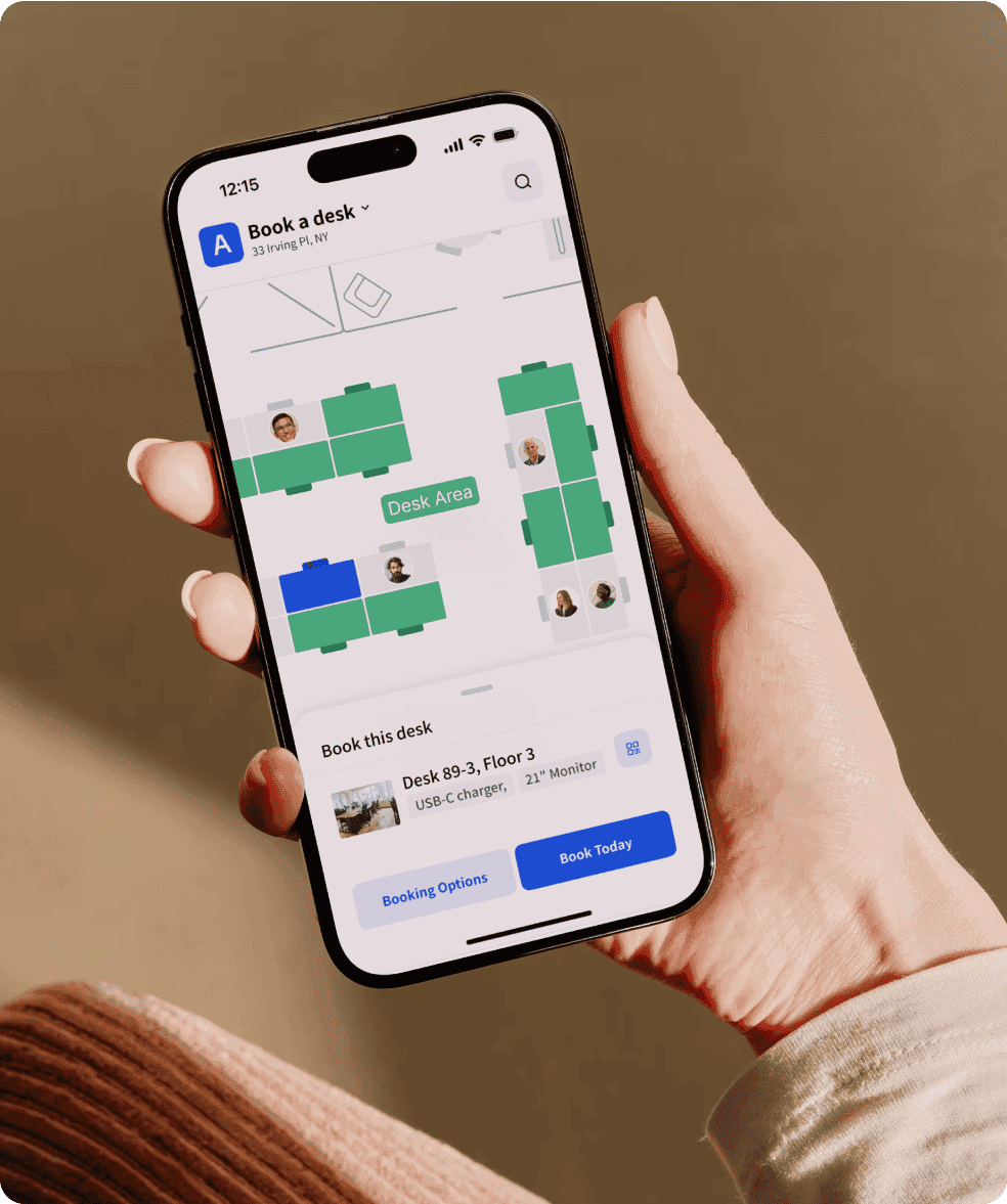 A hand holding a smartphone displaying a desk booking app, showing a floor plan with available desks and an option to book a specific desk.